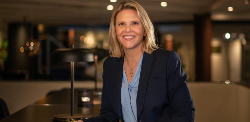 Sylvi Listhaug, smilende i blå dressjakke og lyseblå overdel. Lener seg svakt fremover mot et bord. Uskarp bakgrunn. Foto.