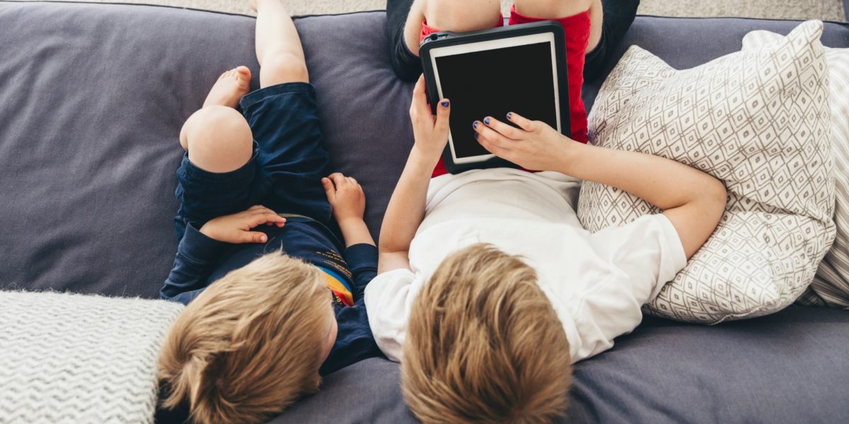 To barn sitter i en sofa. Den ene har holder en ipad i hendene som begge ser på. Foto.